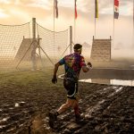 OCR athlete racing across muddy terrain with international flags at an obstacle course event