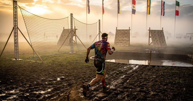 OCR athlete racing across muddy terrain with international flags at an obstacle course event
