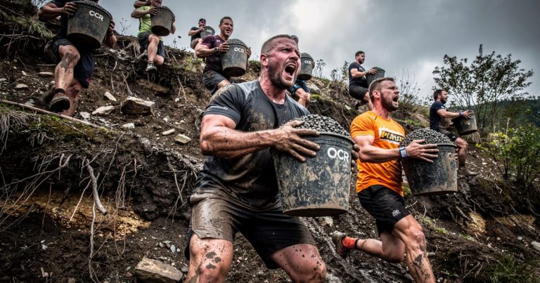 OCR athletes carrying heavy gravel buckets up a steep muddy hill during a race