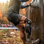 Adaptive OCR athlete scaling a muddy wall obstacle on a dramatic race course