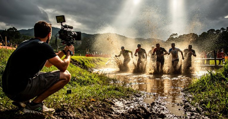Videographer filming OCR athletes charging through a water obstacle on a muddy course